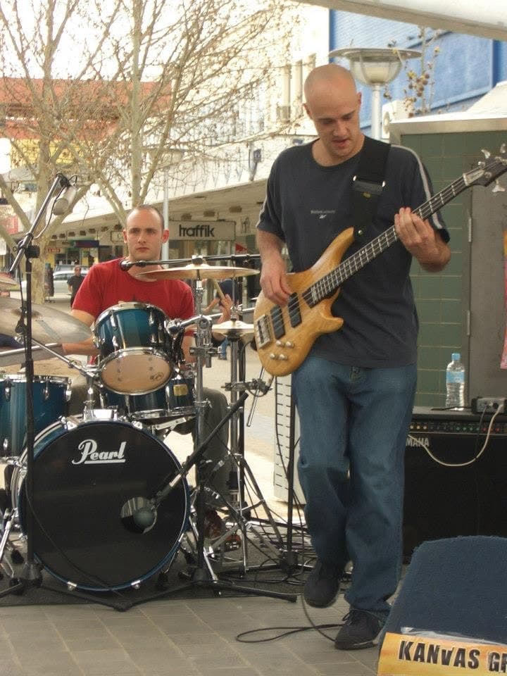 Daytime street show: drummer in a red T-shirt behind a blue Pearl kit and the bassist with a natural-wood bass in the foreground, shopfronts and bare trees behind, a Kanvas Grey banner at the stage edge.