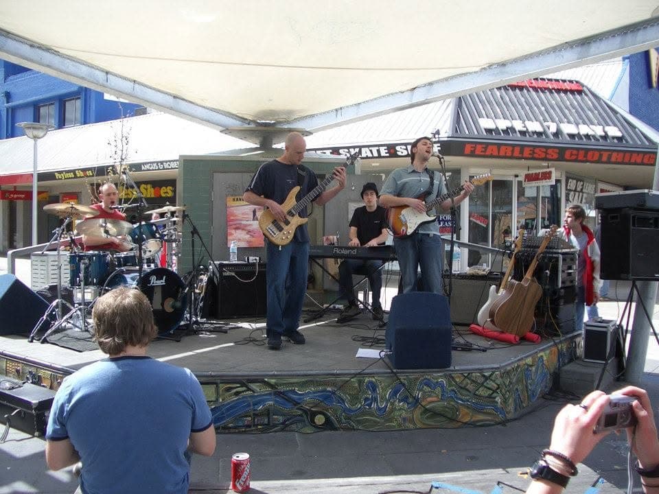 Playing an outdoor mall stage under a white shade sail: drums, bass, a Roland keyboard and the singer with a Stratocaster, shopfronts behind, a mosaic along the stage front and onlookers on the paving.