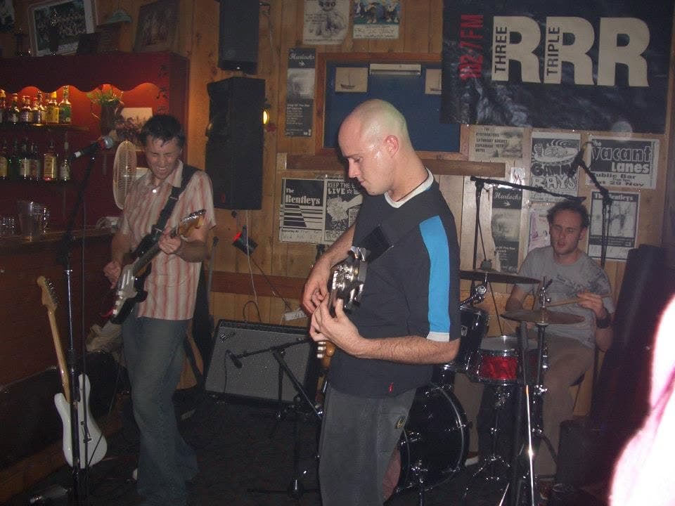 A pub gig beneath a wall of gig posters and a Triple R banner: guitarist in a striped shirt on the left, bassist head-down in the centre, drummer behind a red kit on the right.